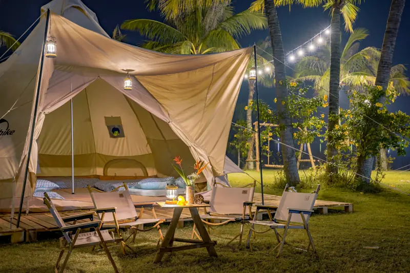 Lều glamping màu be có mái che, bàn ghế gỗ và đèn dây dưới hàng dừa lúc tối, By The Lake Glamping