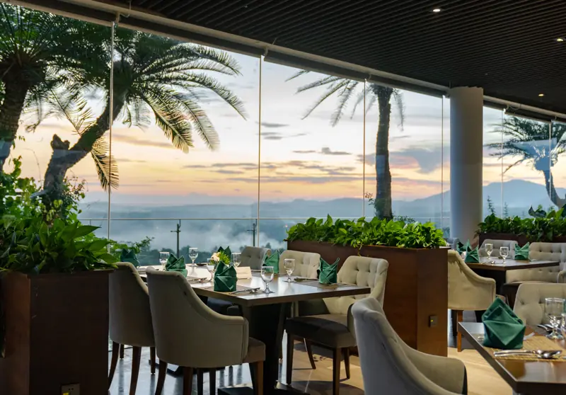 Bàn ăn trải khăn xanh cạnh vách kính Sky Restaurant, bên ngoài là hàng cọ và trời chiều trên hồ