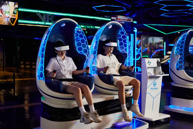 Hai bạn trẻ đeo kính thực tế ảo ngồi trên ghế VR hình quả trứng phát sáng xanh trong khu VR Game