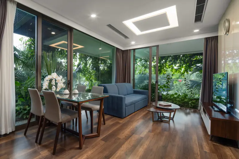 Phòng khách căn Deluxe Sky Residence 1 phòng ngủ: sofa xanh, bàn ăn 4 ghế, cửa kính nhìn ra cây xanh