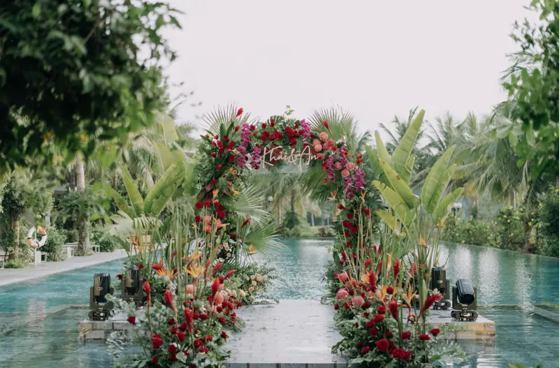 Cổng hoa đỏ và lối đi bắc qua mặt bể bơi giữa vườn cây nhiệt đới ở Flamingo Đại Lải